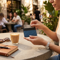 A close-up lifestyle shot of a person holding a slim black card holder wallet with a blue and black checkered pattern, containing a credit card. The person is seated at a stone table in a sunny, outdoor cafe setting with a coffee cup, sunglasses, and a notebook nearby.