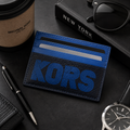 A blue and black leather card holder wallet with a large embossed 'KORS' logo, styled on a dark desk with a coffee cup, pen, sunglasses, notebook, and wristwatch.