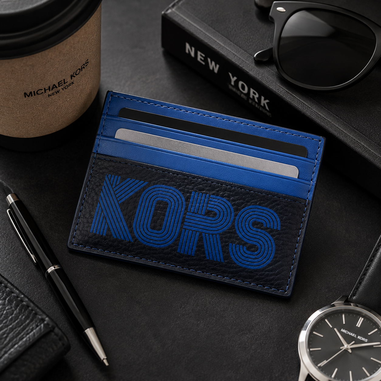 A blue and black leather card holder wallet with a large embossed 'KORS' logo, styled on a dark desk with a coffee cup, pen, sunglasses, notebook, and wristwatch.