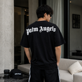 A man viewed from behind wearing a black Palm Angels t-shirt with the brand name in white gothic lettering across the back, paired with black track pants with white side stripes. A matching black Palm Angels cap and a coffee cup sit on a table in the foreground of a modern living room setting.