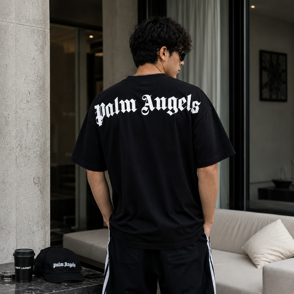 A man viewed from behind wearing a black Palm Angels t-shirt with the brand name in white gothic lettering across the back, paired with black track pants with white side stripes. A matching black Palm Angels cap and a coffee cup sit on a table in the foreground of a modern living room setting.