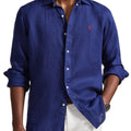 A man wearing a blue long-sleeve linen button-down shirt with rolled-up sleeves and white pants, shown from the neck down against a white background.