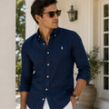 A man wearing a navy blue long-sleeve button-down shirt with a small white embroidered polo player logo on the chest, styled with white pants and sunglasses, standing in front of a white house porch.
