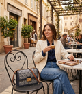 A woman sits at an outdoor cafe table holding a coffee cup, wearing a cream blazer, navy top, and blue jeans. A patterned navy and beige handbag with brown leather straps rests on the chair beside her.