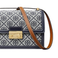 A close-up of a handbag featuring a navy and white monogram pattern, a navy leather trim, a gold-tone turn-lock clasp, and a brown leather shoulder strap on a white background.