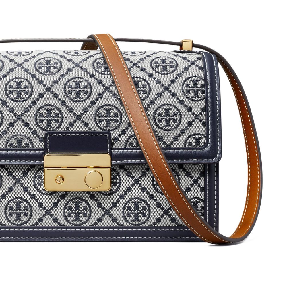 A close-up of a handbag featuring a navy and white monogram pattern, a navy leather trim, a gold-tone turn-lock clasp, and a brown leather shoulder strap on a white background.