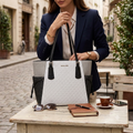 A woman in a navy blazer holds a Michael Kors tote bag with a white monogram pattern, black leather handles, and gray side panels, set in an outdoor cafe environment with a notebook, sunglasses, and a coffee cup on a table.